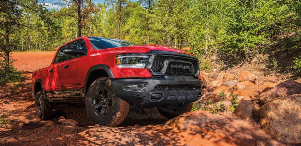 A red Ram 1500 truck navigates rocky terrain surrounded by trees and a clear blue sky.