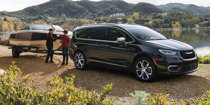 2026 Chrysler Pacifica minivan towing a camper by a lakeside with people nearby