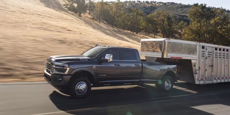 Side view of a heavy-duty pickup truck towing a large enclosed livestock trailer on a highway, with dry hills and trees in the background.