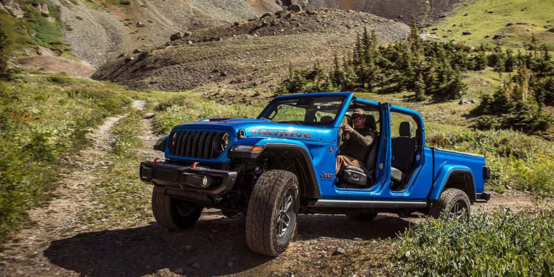 Blue Jeep-style off-road vehicle driving on a rugged mountain trail with the doors removed, showing a driver inside and surrounding green hillside terrain.