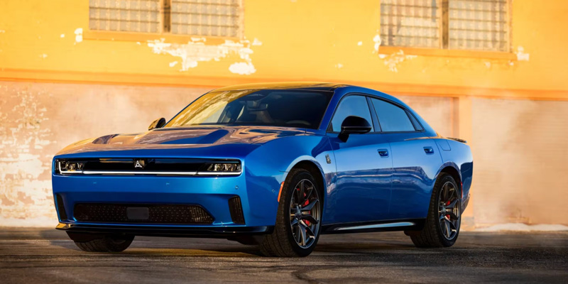 Front three-quarter view of a blue Dodge Charger parked in front of a building with warm sunlight reflecting off its body, highlighting its sporty design and performance stance.
