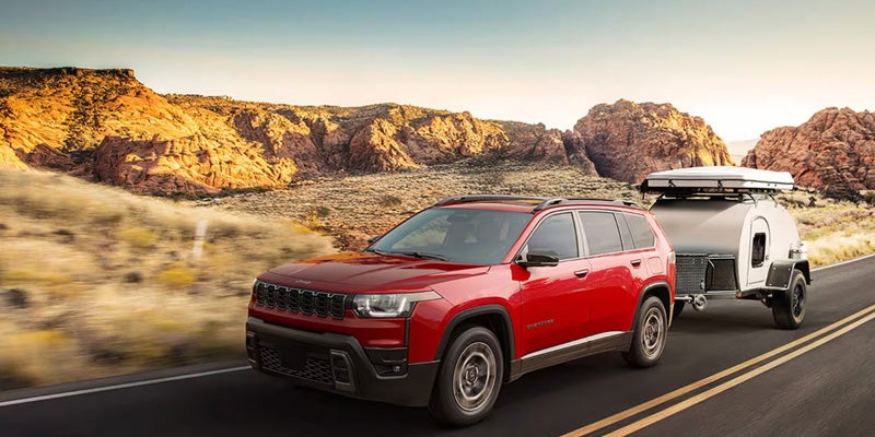 Red Jeep Grand Cherokee towing a small camper trailer along a desert highway with rocky cliffs in the background.